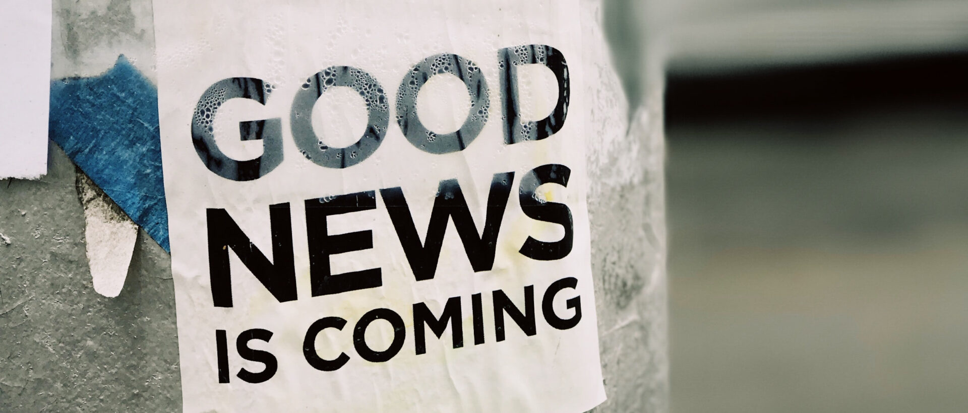 An einer Plakatsäule hängt ein Zettel, auf dem in grossen, schwarzen Buchstaben "Good News is Coming" gedruckt ist.