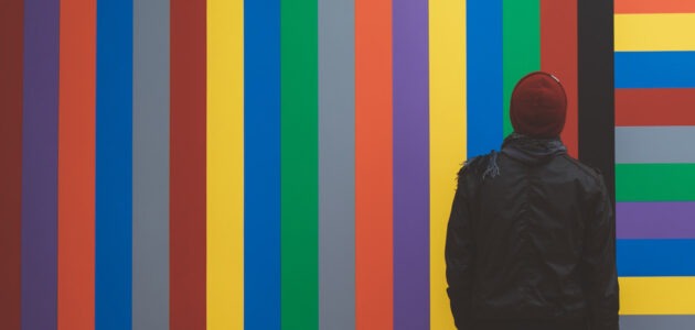Eine Person in dunkler Kleidung steht vor einer Wand mit Farbstreifen. Die Farbstreifen können als Symbolik für Design oder Designsystem verstanden werden