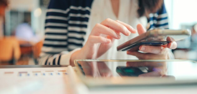Eine Person bedient ein Smartphone über einem Tablet auf einem Tisch. Im Vordergrund liegen Unterlagen mit Diagrammen und Auswertungen. Die Szene verbindet mobile Mediennutzung mit Alltag und digitalem Verhalten von Generation Z und Millennials.