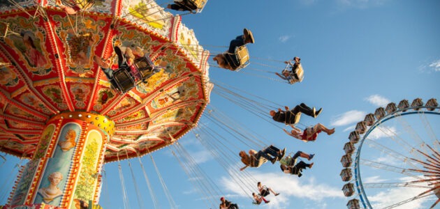 Buntes Kettenkarussell mit Menschen in fliegenden Sitzen vor blauem Himmel und einem Riesenrad. Das Motiv symbolisiert ein Karussell auf Websites.