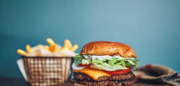Ein Hamburger mit Fleisch, Käse, Tomaten, Salat und Sauce liegt auf einem Holzbrett, dahinter steht eine Portion Pommes frites. Das Motiv spielt auf das Hamburger-Menü für die mobile Navigation an.