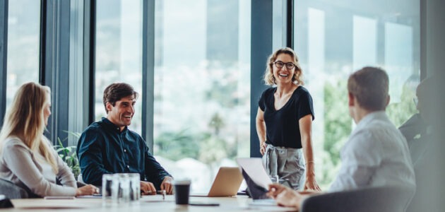 Vier Mitarbeitende tauschen sich während einer Besprechung in einem hellen Büro aus. Eine Frau steht lachend am Tisch und spricht zur Gruppe. Das Bild symbolisiert Corporate Influencer und Employee Advocacy.