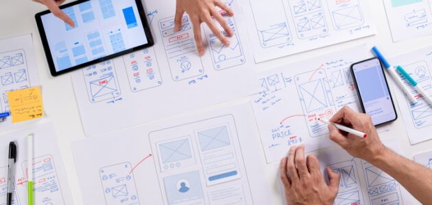 Mehrere Personen arbeiten gemeinsam an handgezeichneten Wireframes für Websites und mobile Apps. Auf dem Tisch liegen Papierentwürfe, ein Tablet und ein Smartphone zur Planung einer Informationsarchitektur.