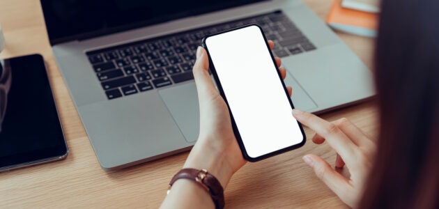 Eine Person hält ein Smartphone mit leerem weissem Bildschirm vor einem geöffneten Laptop und tippt auf das Display. Das Bild steht für das Simulieren und Testen einer mobilen Website-Ansicht auf dem Desktop.