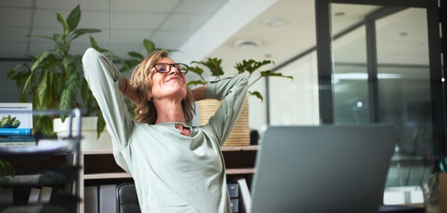 Eine Frau sitzt entspannt mit hinter dem Kopf verschränkten Armen vor einem Laptop in einem hellen Büro. Das Bild steht für die Nutzung neuer Profilfunktionen auf LinkedIn.