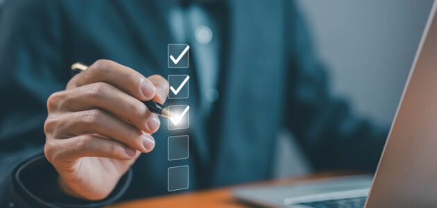 Eine Person markiert mit einem Stift Punkte auf einer digitalen Checkliste neben einem Laptop. Das Bild symbolisiert die Prüfung einer Benutzeroberfläche anhand der Usability-Heuristiken nach Jakob Nielsen.
